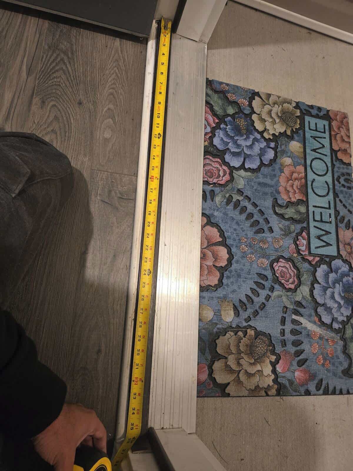 Un mètre ruban posé contre une porte, avec un paillasson orné de motifs floraux sur lequel est écrit « BIENVENUE ».