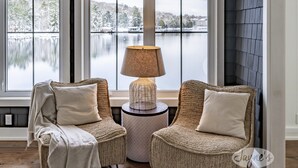 Two wicker chairs with cushions, a round table with a lamp, and a view of a lake through large windows.