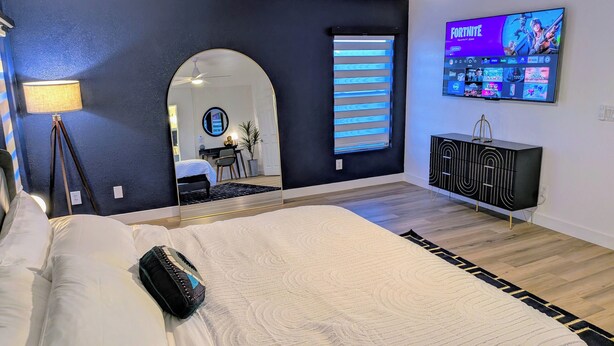 A modern bedroom with a large bed, a TV mounted on the wall, a mirror, and a desk with a chair. - 8 BEDROOM Fun Smart Stylish Home, 8 Minutes to the Strip (Las Vegas)