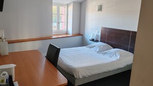 A hotel room with a bed, a desk, a chair, a window, and a nightstand with a lamp. - O'Fun Art  (Airvault)