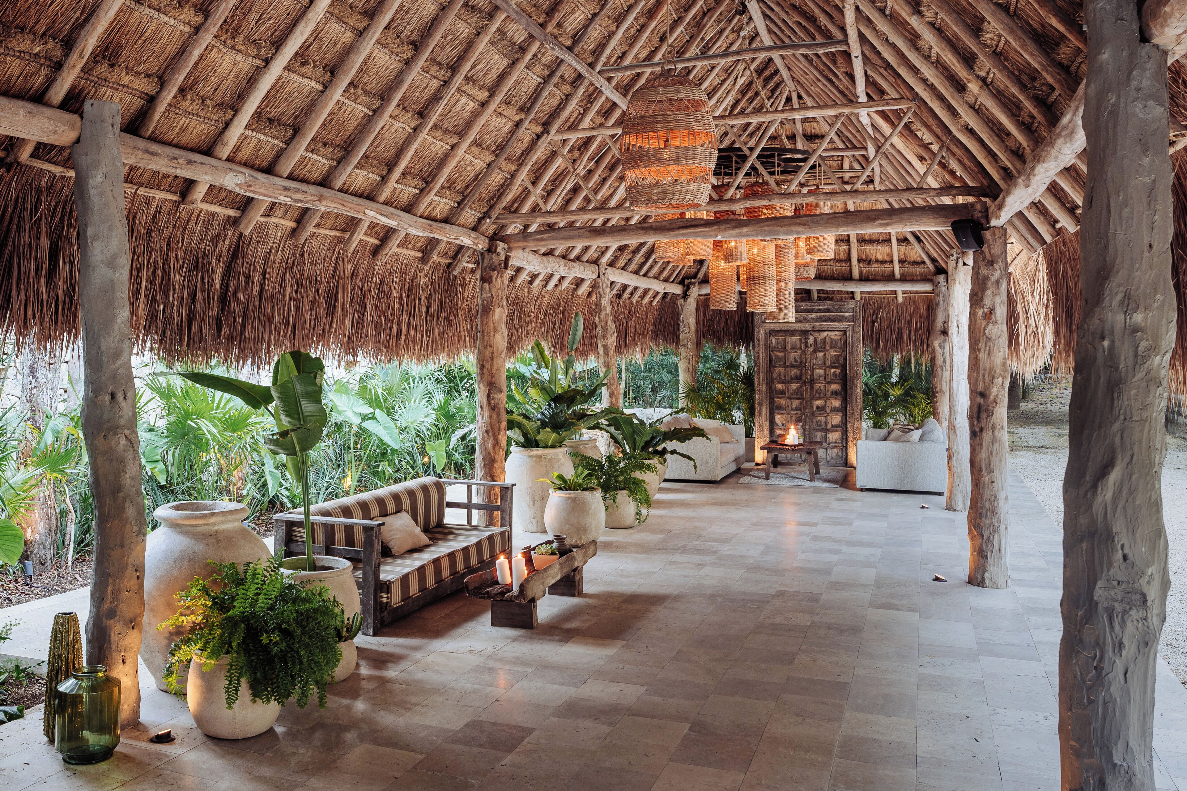 A covered outdoor lounge area with wooden beams and thatched roof, furnished with sofas and planters.