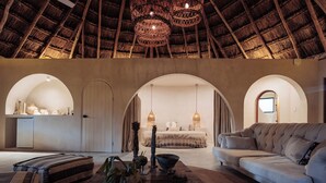 Een ruime kamer met een hoog rieten plafond, een grote bank, een bed en een kleine kitchenette.