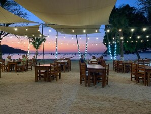 An outdoor dining area with tables and chairs, string lights, and a view of the beach and ocean at sunset. - Sea Bird #2 1 Bed 2 Bath Charming Loft in Las Palmas (Playas del Coco)