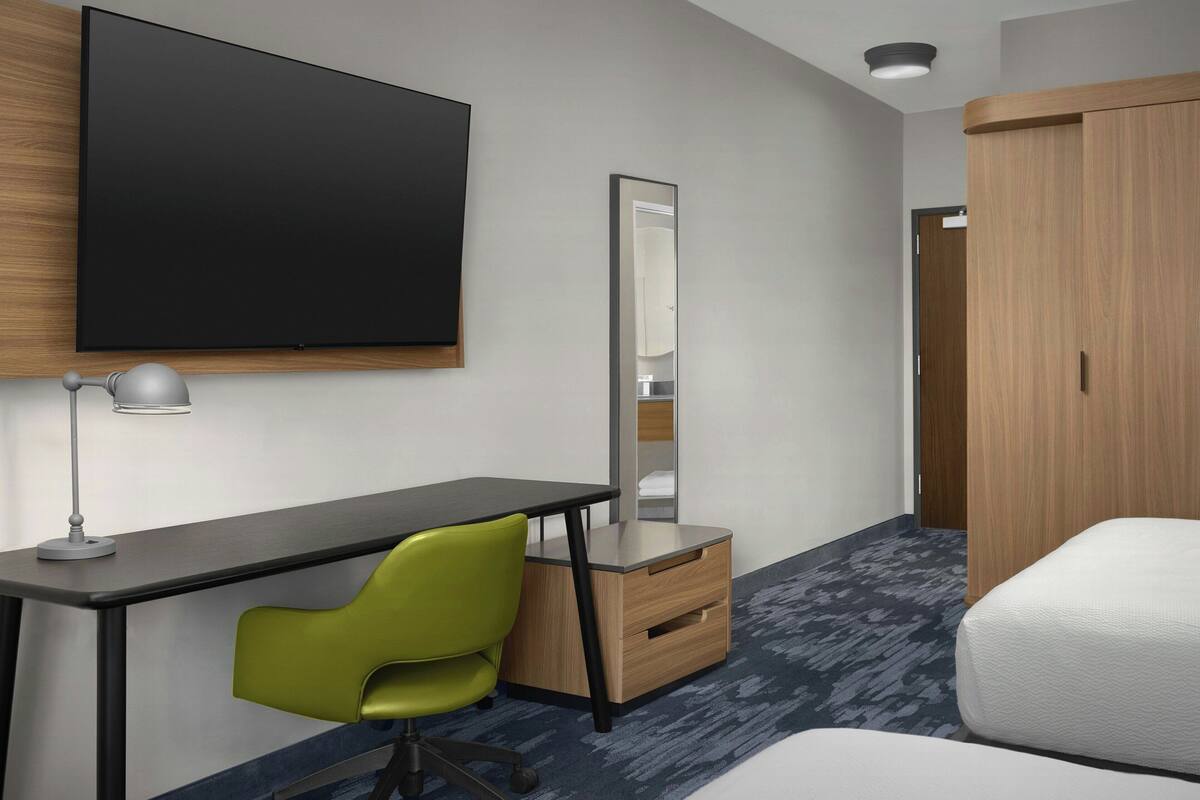 A hotel room with a flat-screen TV, a desk with a chair, a mirror, a wardrobe, and a bed.