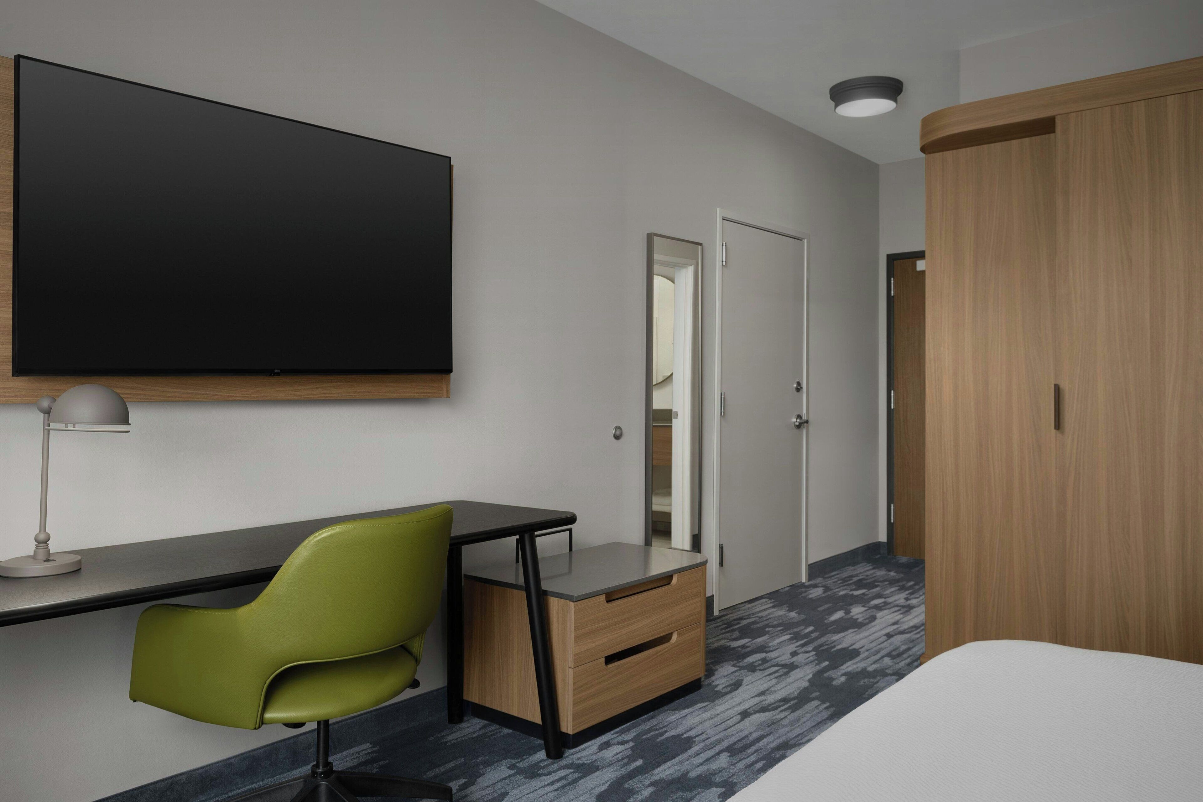 A modern hotel room with a large flat-screen TV mounted on the wall, a desk with a chair, a wardrobe, and a mirror.