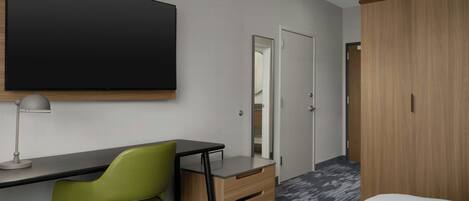 A modern hotel room with a large flat-screen TV mounted on the wall, a desk with a chair, a wardrobe, and a mirror.