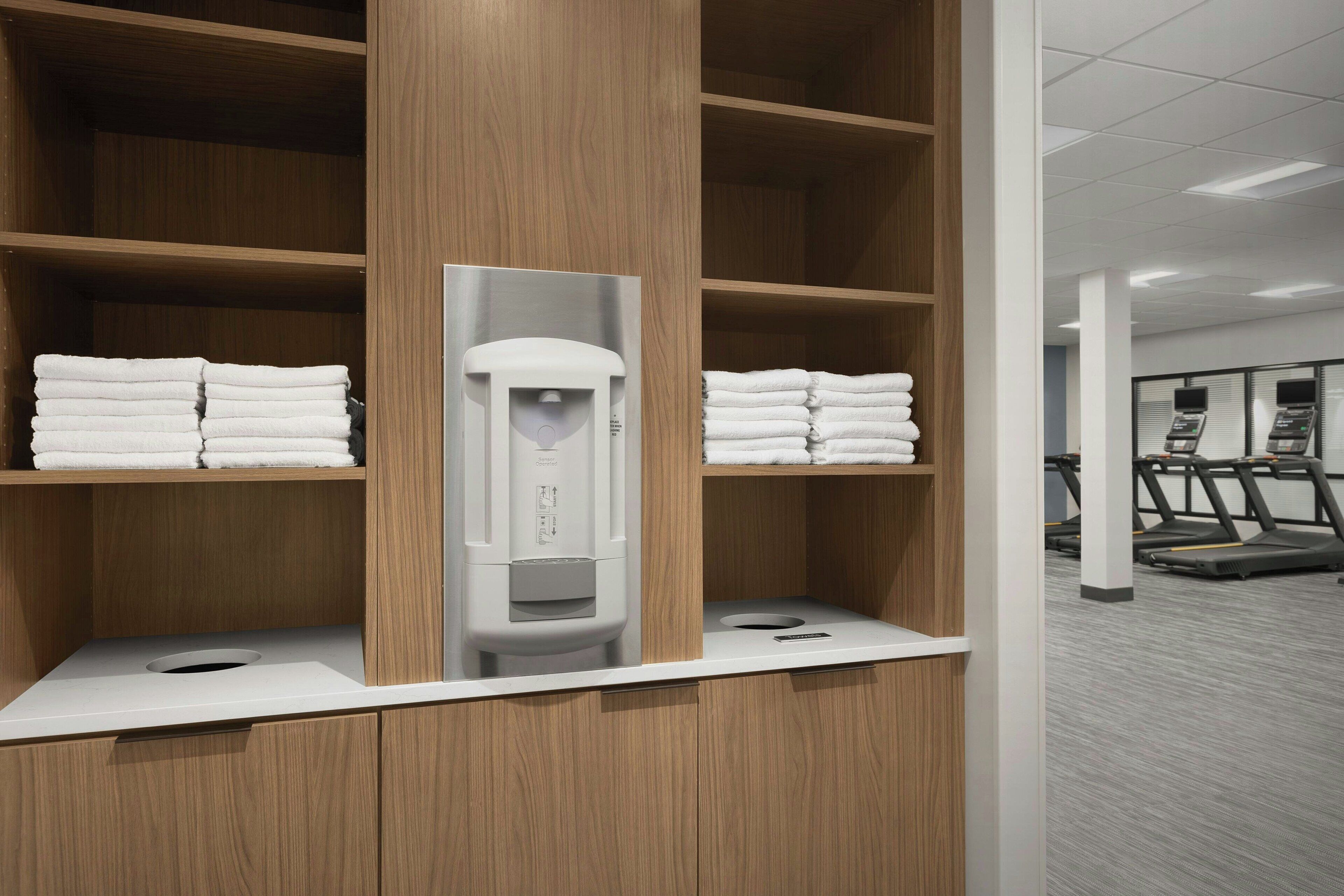 A hotel bathroom with wooden shelves, a towel dispenser, and a treadmill in the background.