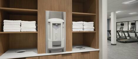 A hotel bathroom with wooden shelves, a towel dispenser, and a treadmill in the background.