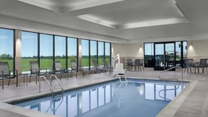 Una piscina interior moderna con paredes de vidrio transparente, un solárium con baldosas y múltiples sillones de descanso.