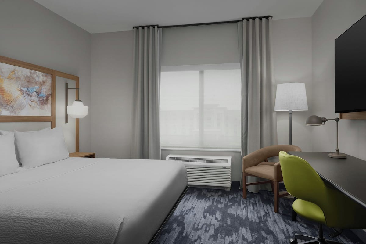 A hotel room with a large bed, a desk with a chair, a flat-screen TV, a lamp, and a window with curtains.