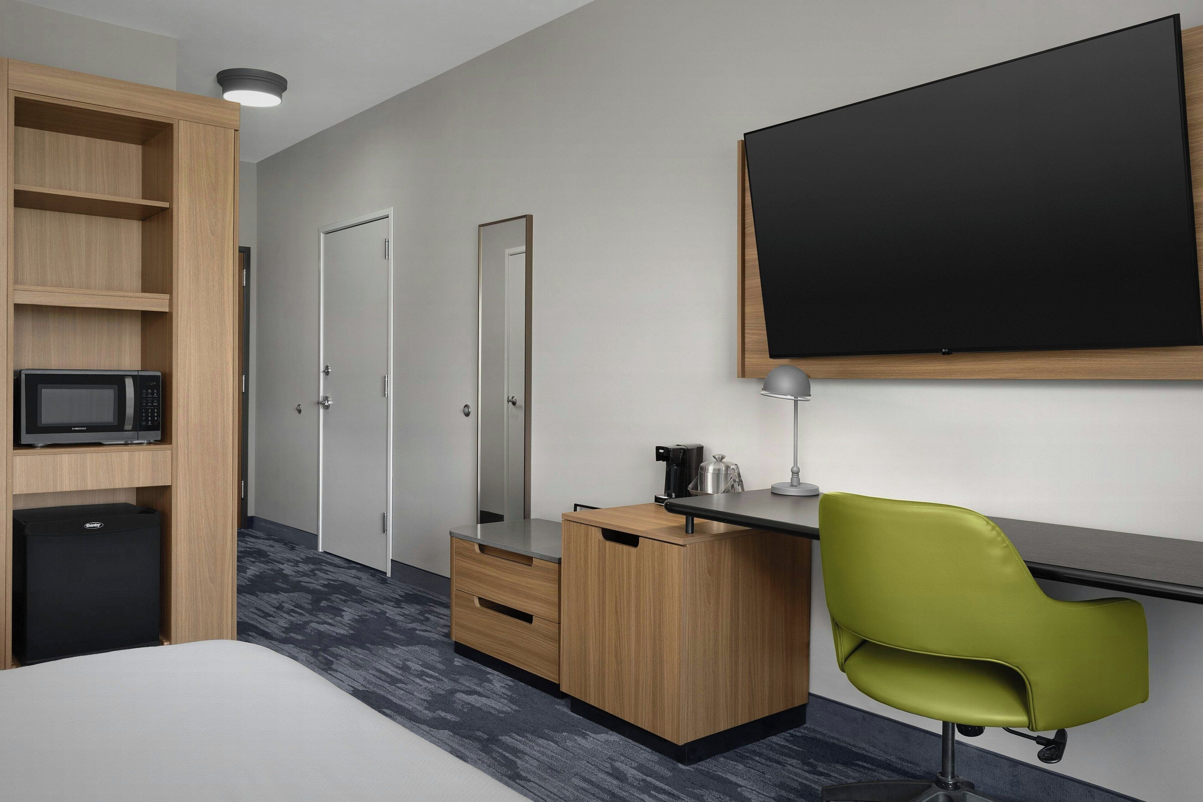 A hotel room with a bed, a desk with a chair, a microwave, a small refrigerator, a mirror, a blackboard, and a wardrobe with shelves.