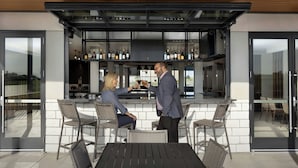 Zona de bar al aire libre con diseño moderno, que incluye una barra, asientos y vistas al interior.