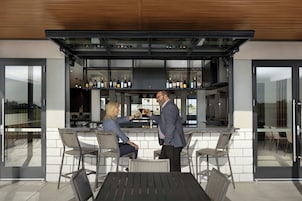 Zona de bar al aire libre con diseño moderno, que incluye una barra, asientos y vistas al interior.