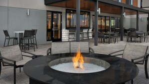 Ein Sitzbereich im Freien mit einem runden Feuerkorb, umgeben von Stühlen, einer überdachten Terrasse und einem Gebäude mit großen Fenstern im Hintergrund.