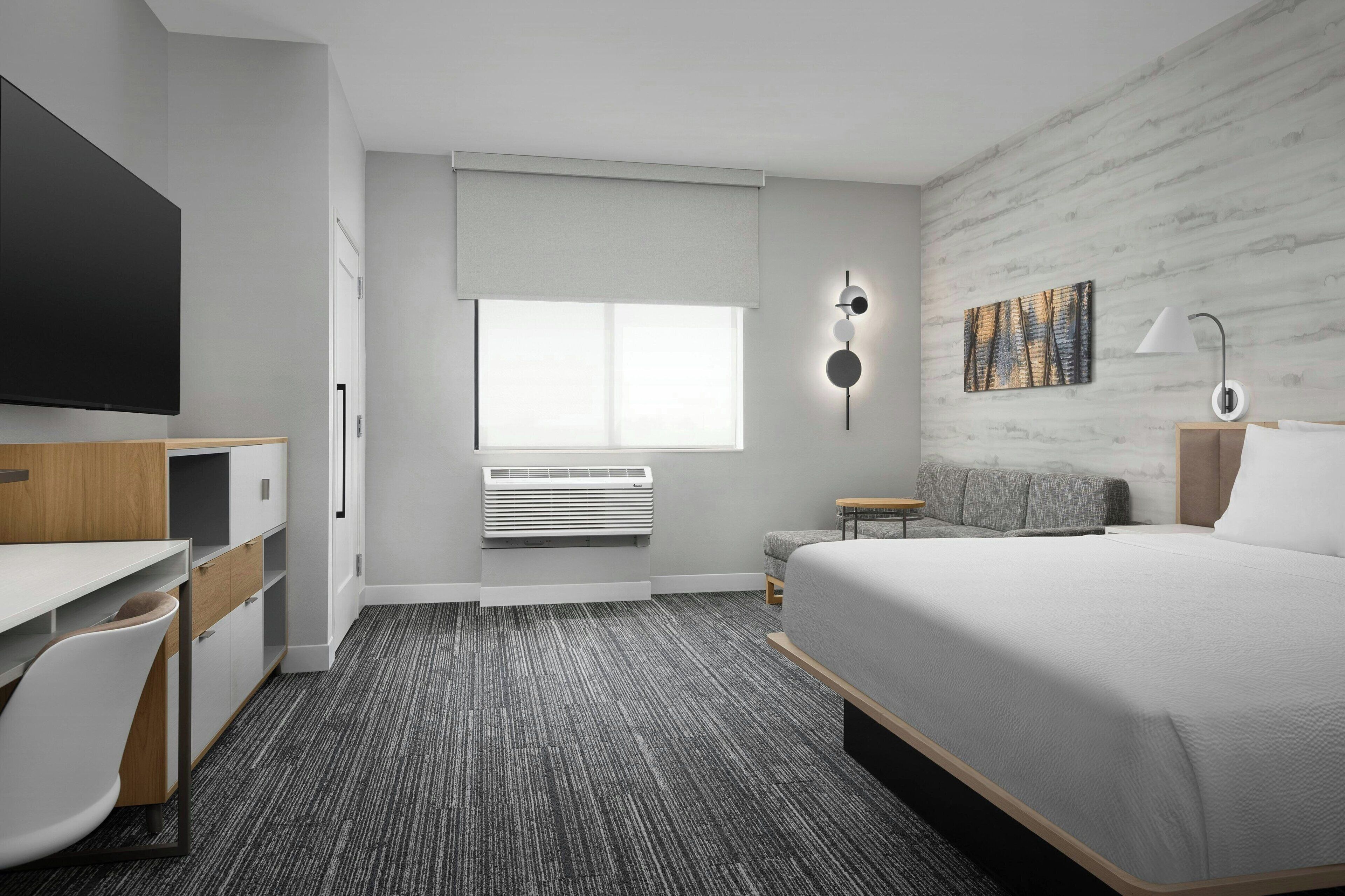 A modern hotel room with a large bed, a desk with a chair, a flat-screen TV, a wall-mounted air conditioner, and a window with blinds.