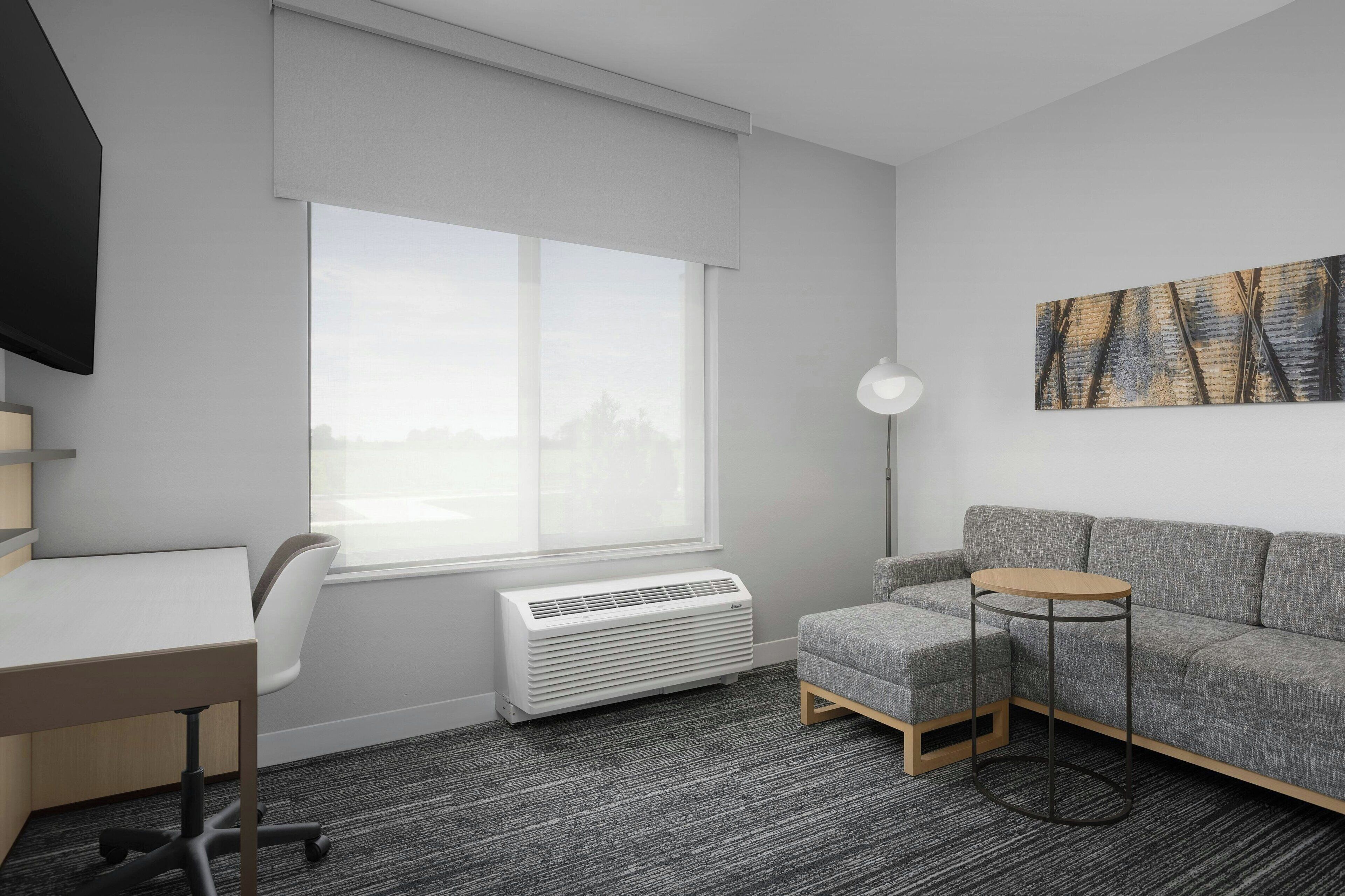A modern room with a desk, a grey sofa, a round table, a standing lamp, and a large window with blinds.