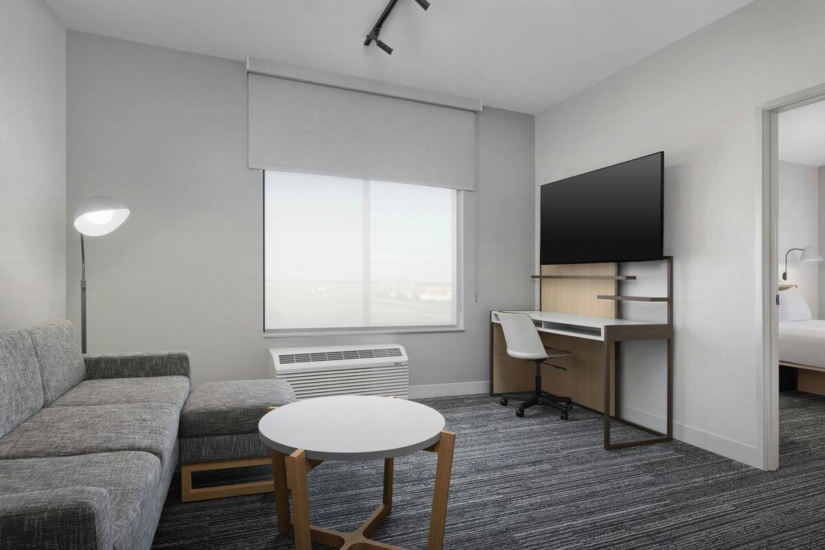 Ein modernes Hotelzimmer mit grauem Sofa, einem runden Tisch, einem Schreibtisch mit Stuhl, einem Flachbildfernseher und einem großen Fenster.