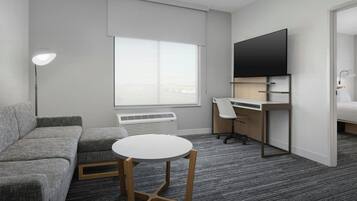 Een moderne hotelkamer met een grijze bank, een ronde tafel, een bureau met stoel, een flatscreen televisie en een groot raam.