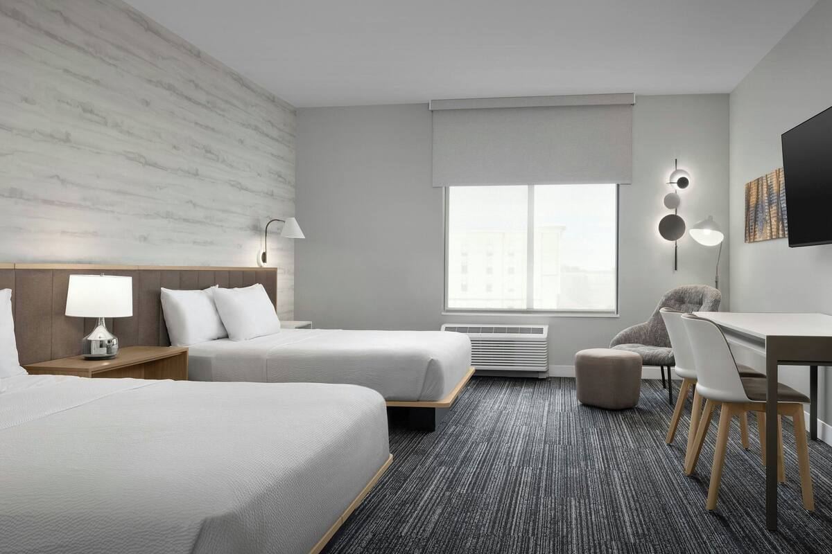 Ein Hotelzimmer mit zwei Betten, einem Schreibtisch, einem Stuhl, einer Lampe, einem Fernseher und einem Fenster mit Blick auf Gebäude.