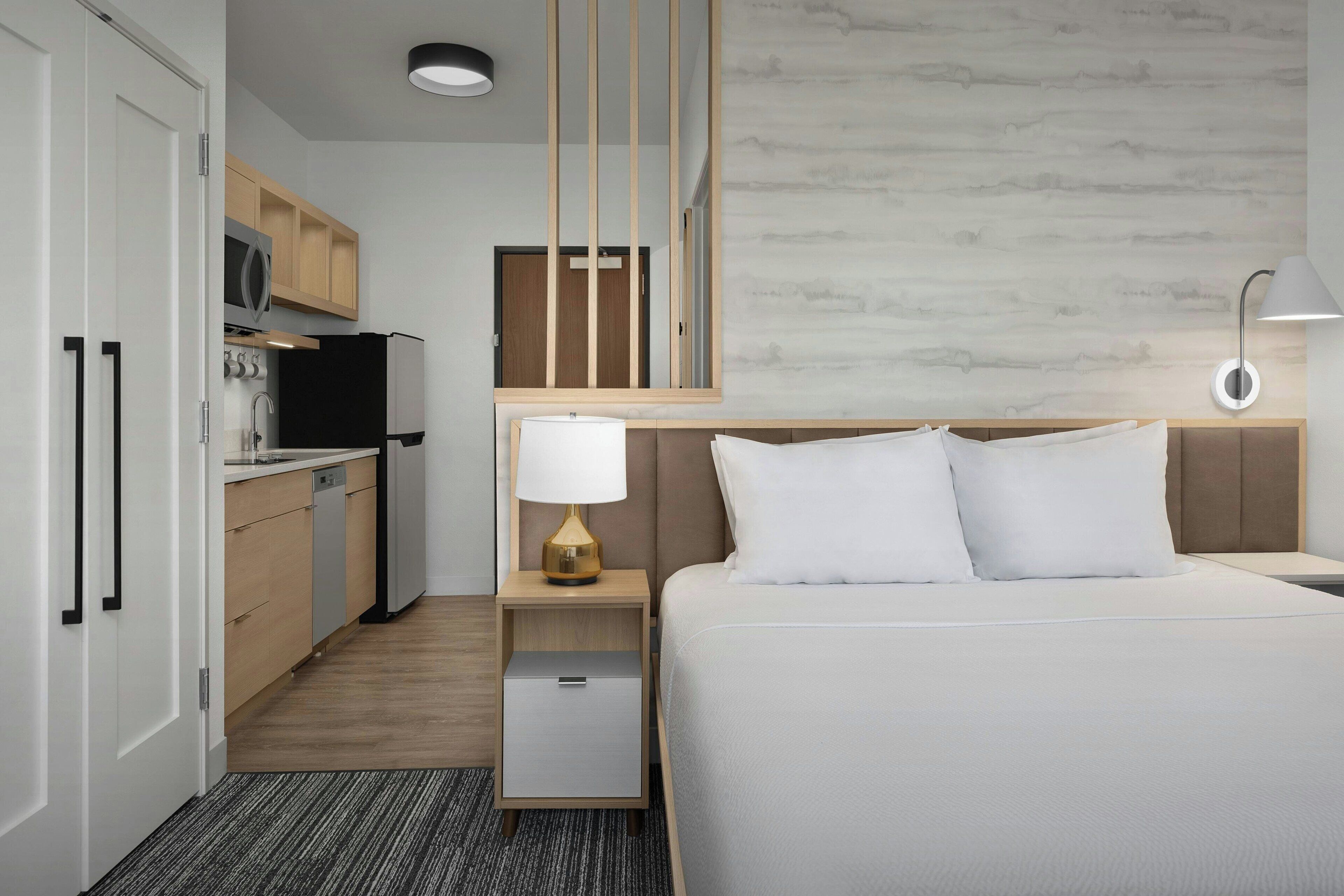 A hotel room with a bed, bedside table, lamp, and a kitchenette with a refrigerator, microwave, and cabinets.