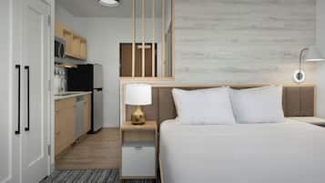A hotel room with a bed, bedside table, lamp, and a kitchenette with a refrigerator, microwave, and cabinets.