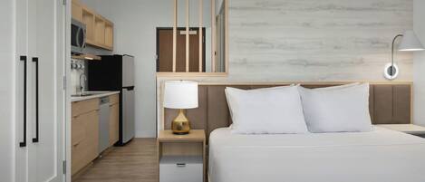 Et hotelværelse med seng, sengebord, lampe og et tekøkken med køleskab, mikroovn og skabe.