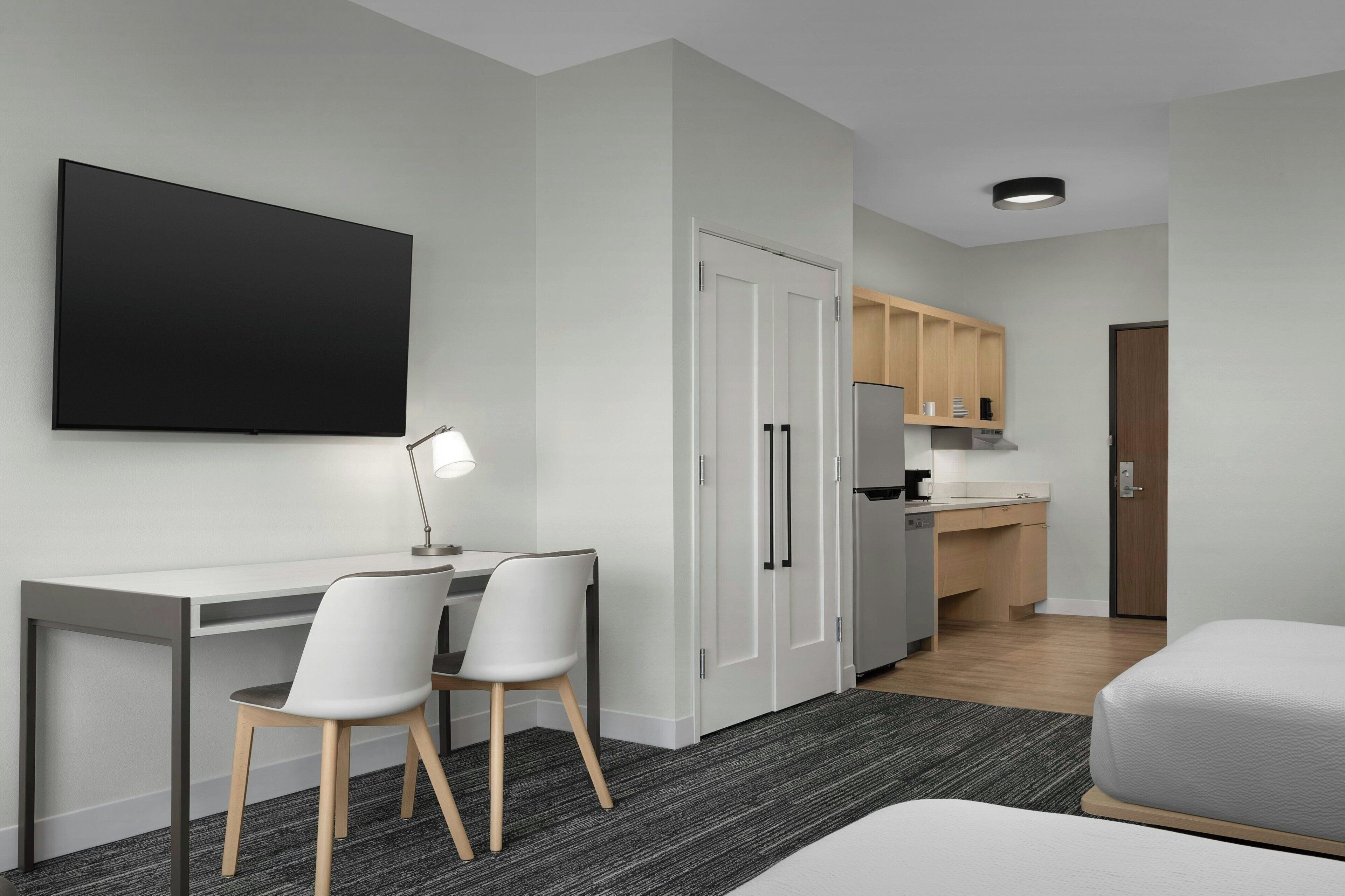 A modern hotel room with a desk, two chairs, a bed, a flat-screen TV, a kitchenette, and a wardrobe.