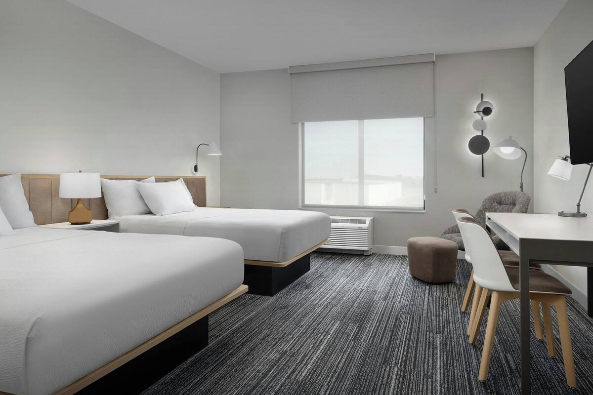 Ein Hotelzimmer mit zwei Betten, einem Schreibtisch, einem Stuhl, einem kleinen Hocker, einem Fenster mit Jalousien und einer Wandlampe.