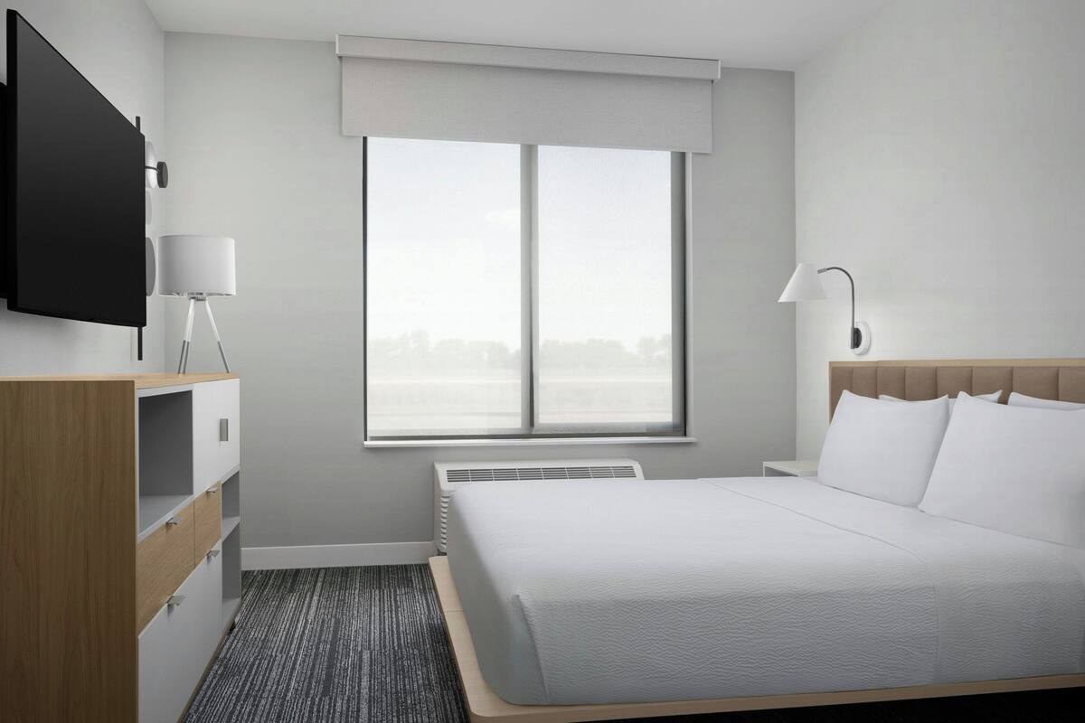 Ein Hotelzimmer mit Bett, Wandfernseher, Nachttisch, Lampe und Fenster mit Blick.