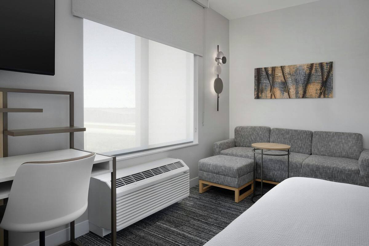 Ein modernes Hotelzimmer mit großem Fenster, grauem Sofa, kleinem runden Tisch, weißem Sessel und einem Bett.