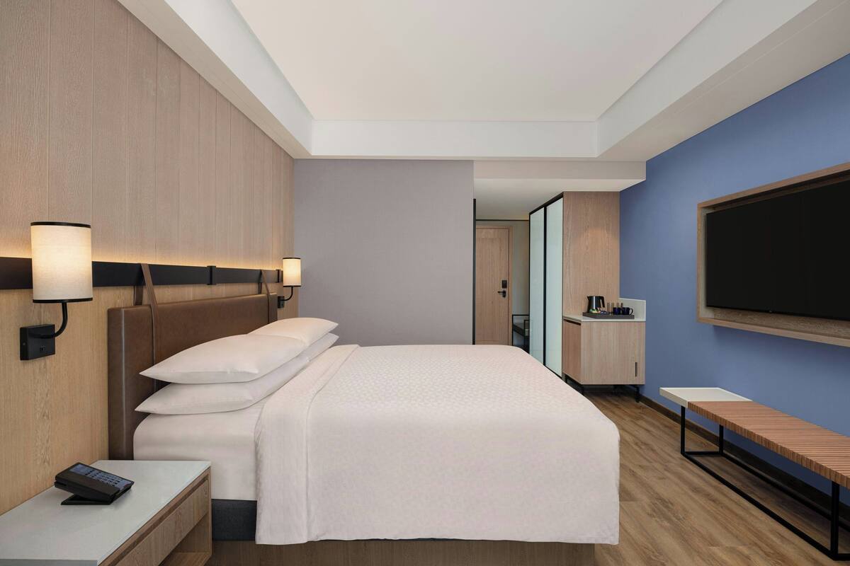 A modern hotel room with a large bed, bedside lamps, a TV, a small desk, and a bench.