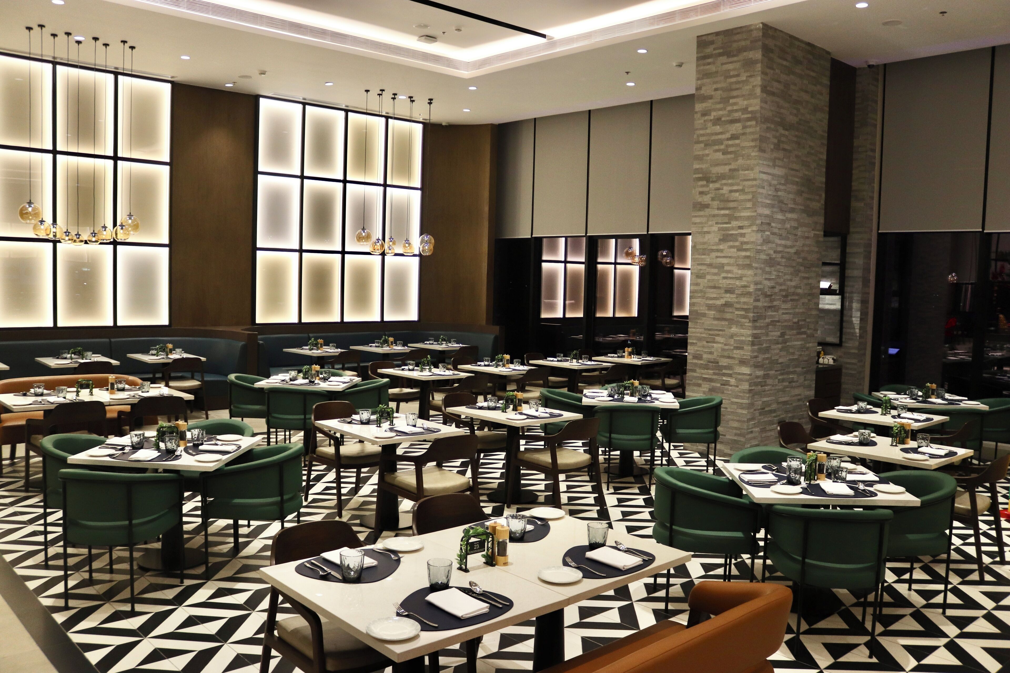 Un restaurante moderno con un piso de patrón geométrico blanco y negro, sillas tapizadas en verde y una zona central de asientos con techo alto e iluminación empotrada.