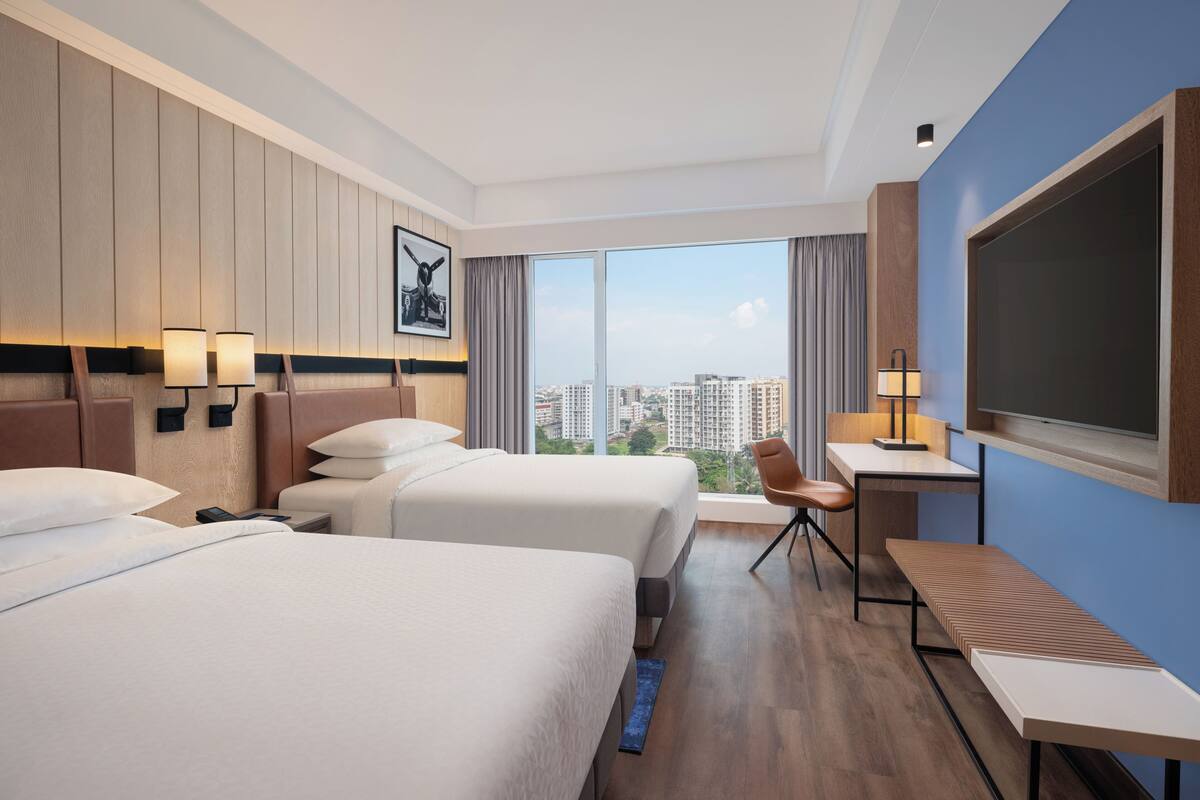A hotel room with two beds, a desk, a chair, a TV, and a large window with a city view.