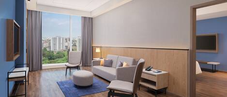A modern hotel room with a large window, a sofa, a chair, a small table, and a TV.