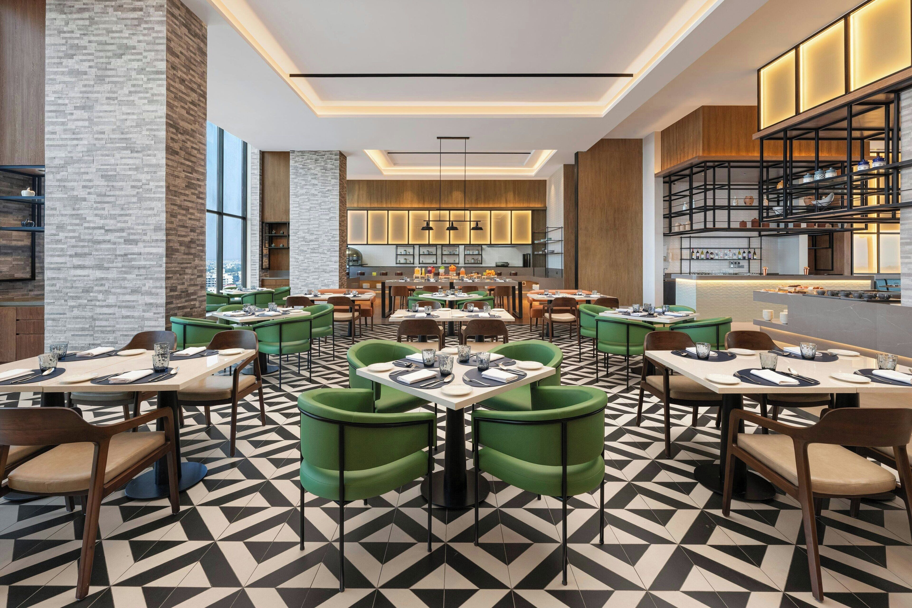 A modern restaurant with multiple dining tables and green chairs, featuring a geometric patterned floor and a kitchen area in the background.