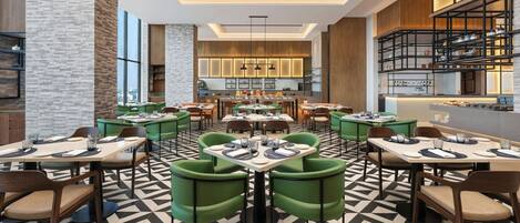 A modern restaurant with multiple dining tables and green chairs, featuring a geometric patterned floor and a kitchen area in the background.