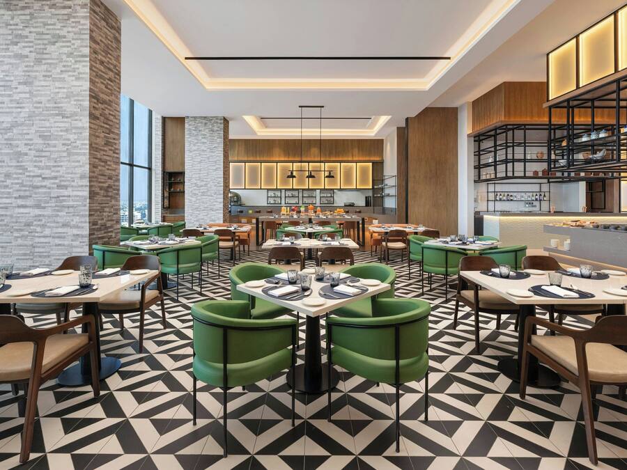 A modern restaurant with multiple dining tables and green chairs, featuring a geometric patterned floor and a kitchen area in the background.