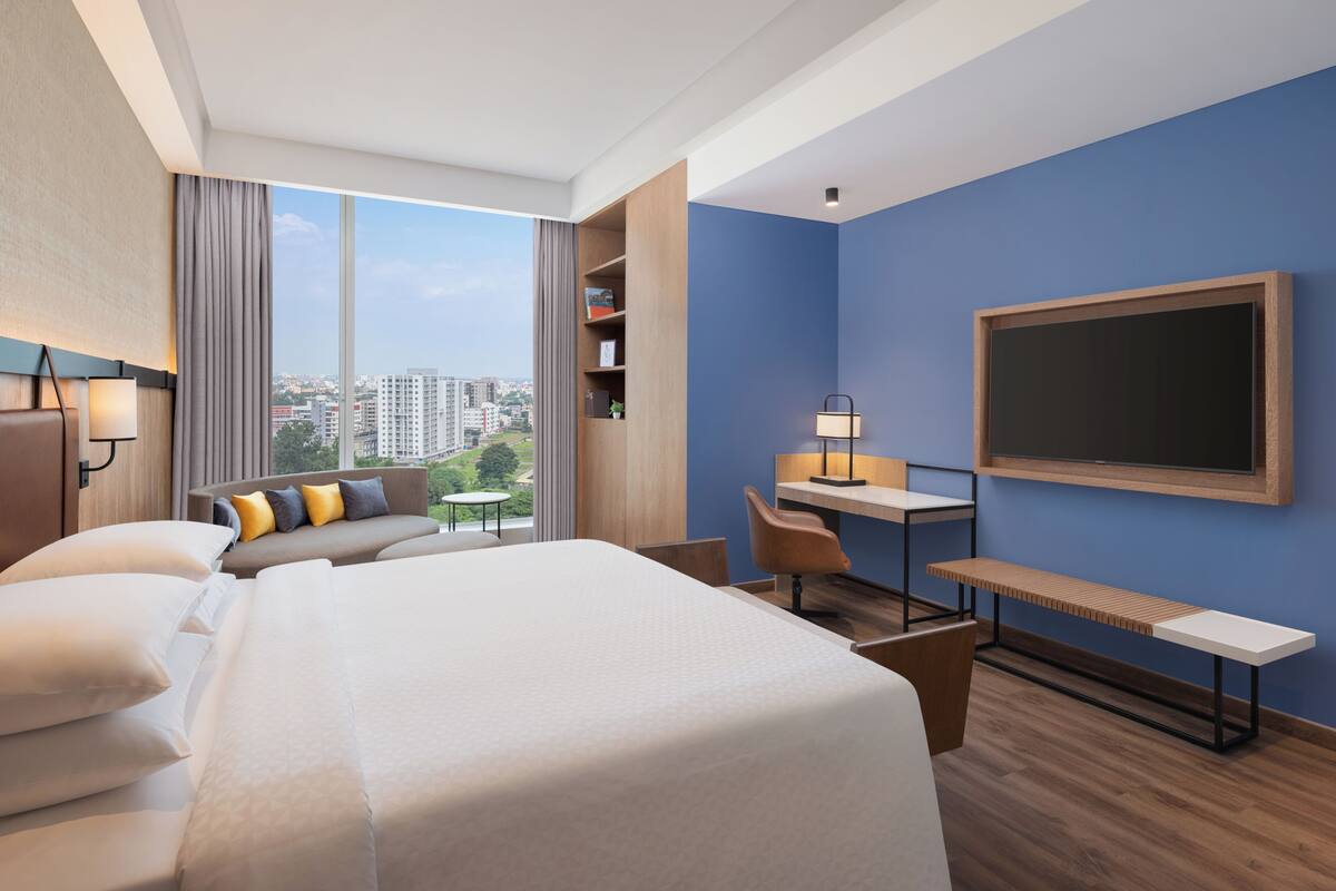 A hotel room with a large bed, a desk, a chair, a bookshelf, and a large window offering a city view.