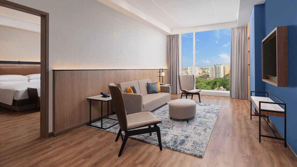 Habitación de hotel con cama, sofá, sillón, televisor y un amplio ventanal con vista a la ciudad.