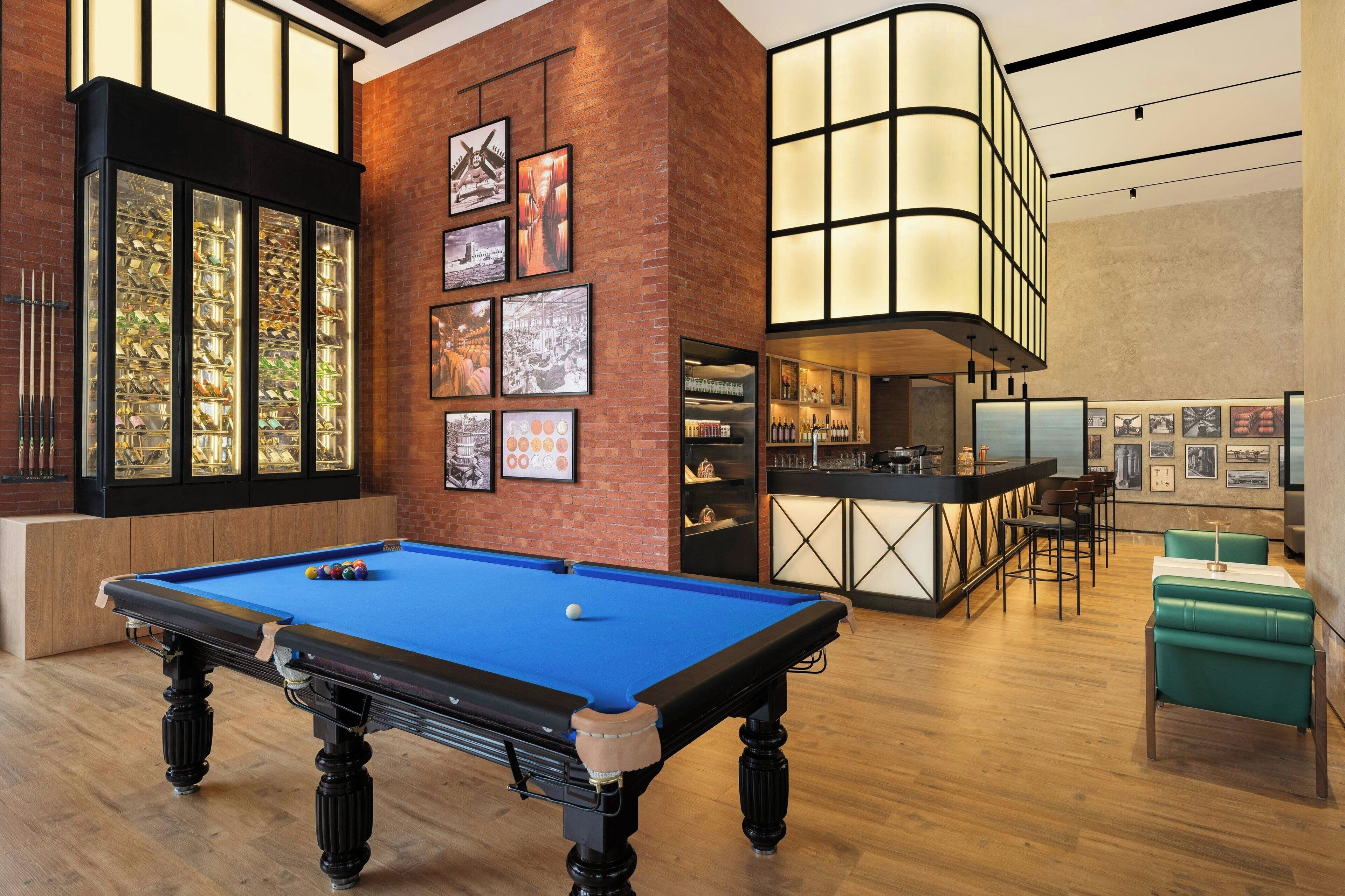 A pool table in a room with a bar area, wooden floors, and framed pictures on the walls.