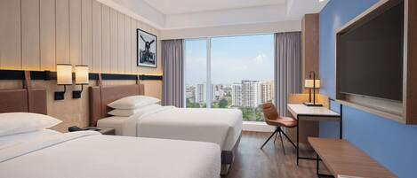 Quarto de hotel com duas camas, uma escrivaninha, uma cadeira, televisão e uma grande janela com vista para a cidade.