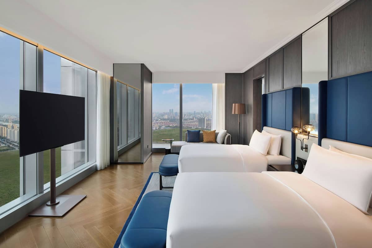 Une chambre d’hôtel moderne dotée de grandes fenêtres, d’un téléviseur à écran plat, de deux lits, d’un coin salon et offrant une vue sur la ville.