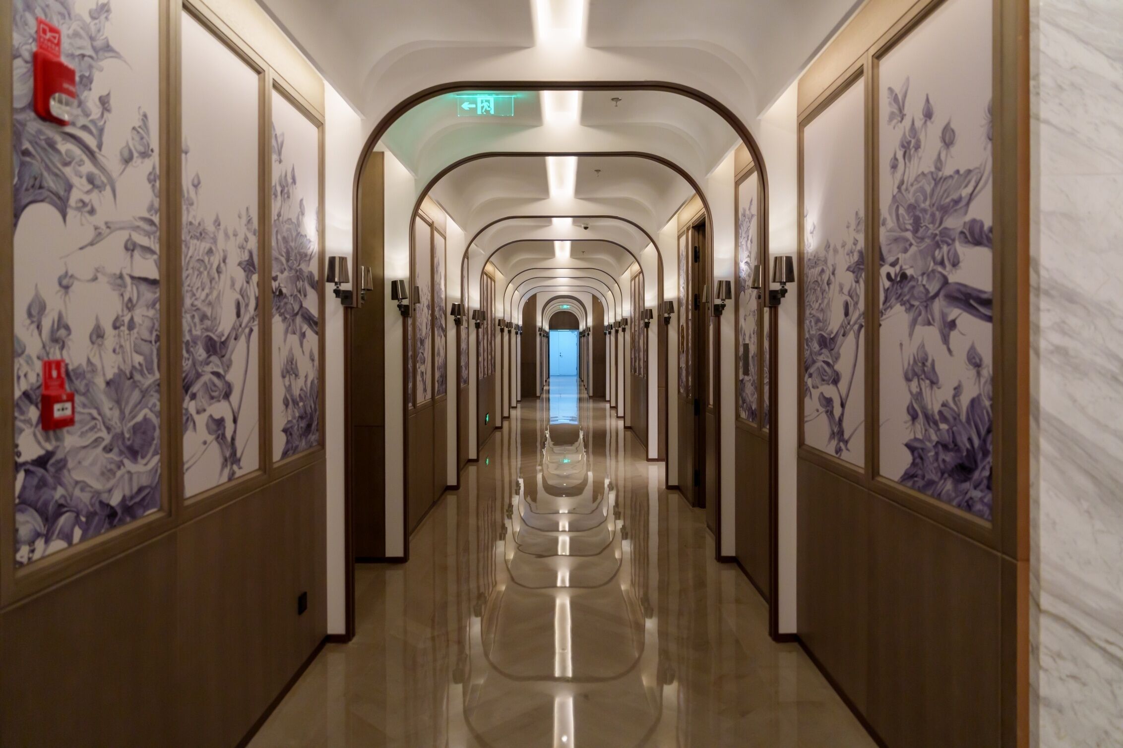 Un largo y estrecho pasillo de hotel con suelo reflectante, flanqueado por paredes con papel tapiz floral, y una puerta de salida azul al final.