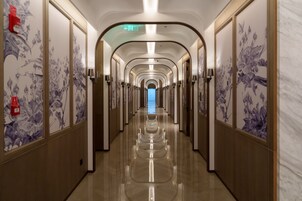 Un largo y estrecho pasillo de hotel con suelo reflectante, flanqueado por paredes con papel tapiz floral, y una puerta de salida azul al final.