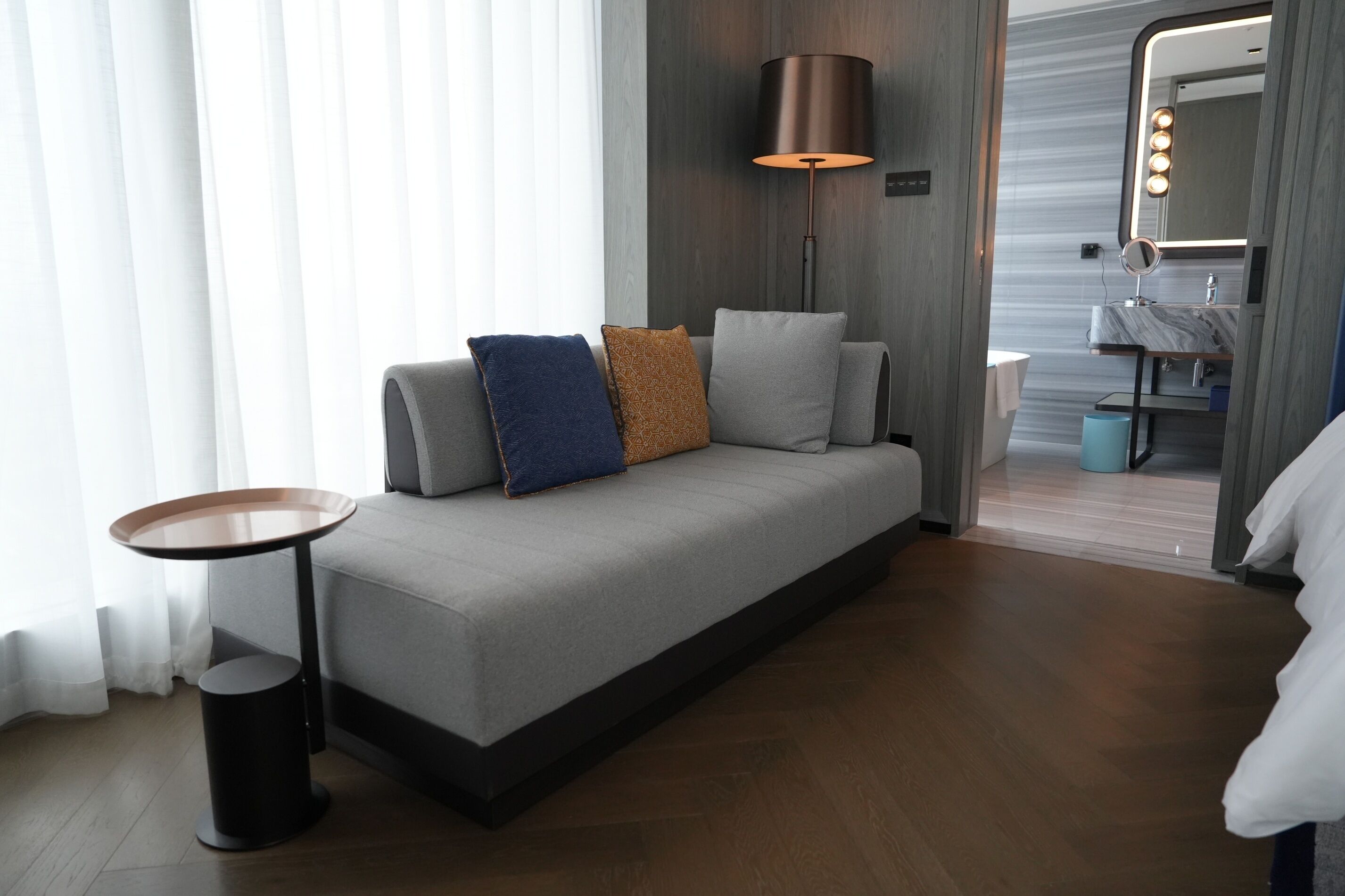 Una habitación de hotel moderna con un sofá gris, una mesita redonda, una lámpara de pie y un baño con tocador y espejo.