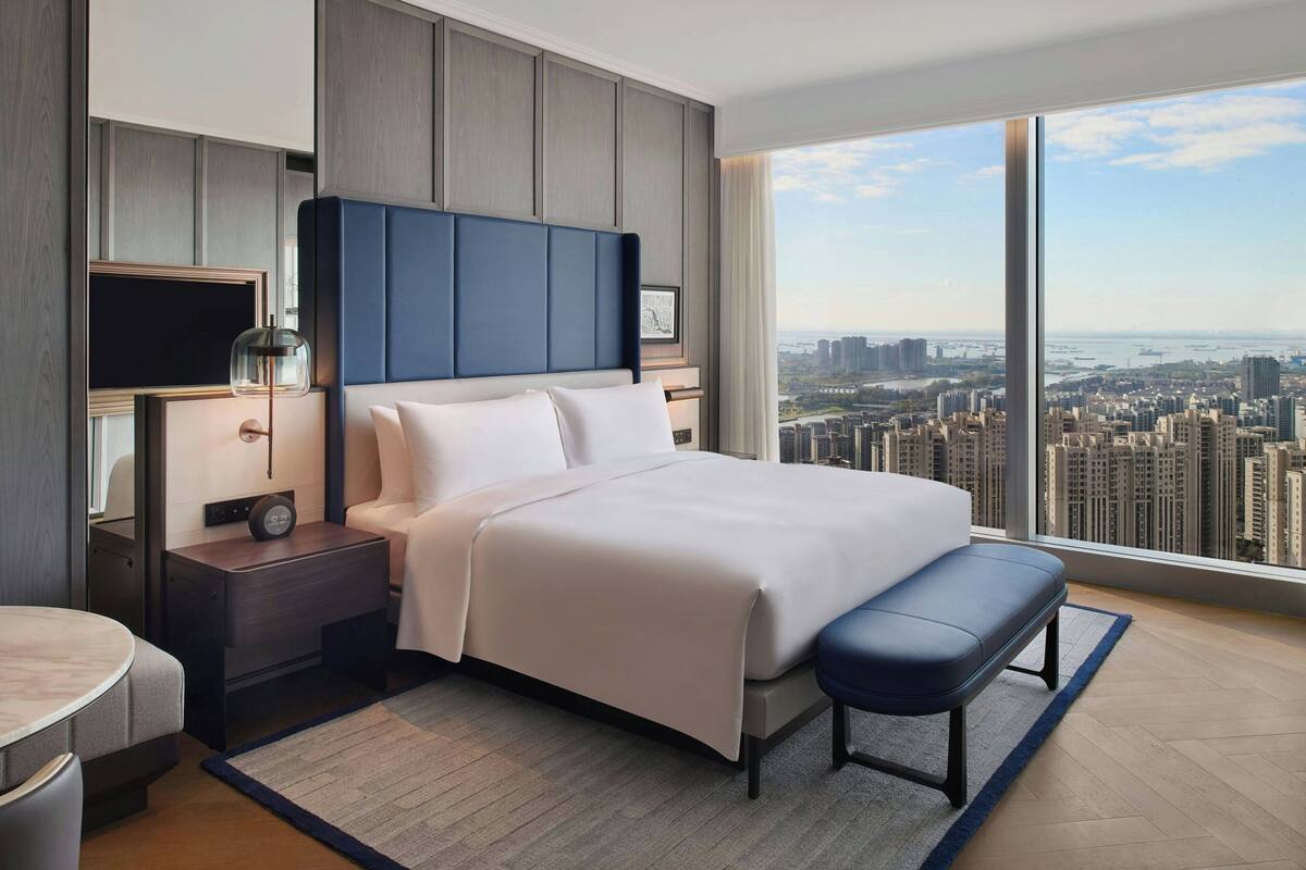 Une chambre d’hôtel moderne avec un grand lit, une table de chevet avec une lampe, une télévision et une vue sur le paysage urbain grâce à de grandes fenêtres.
