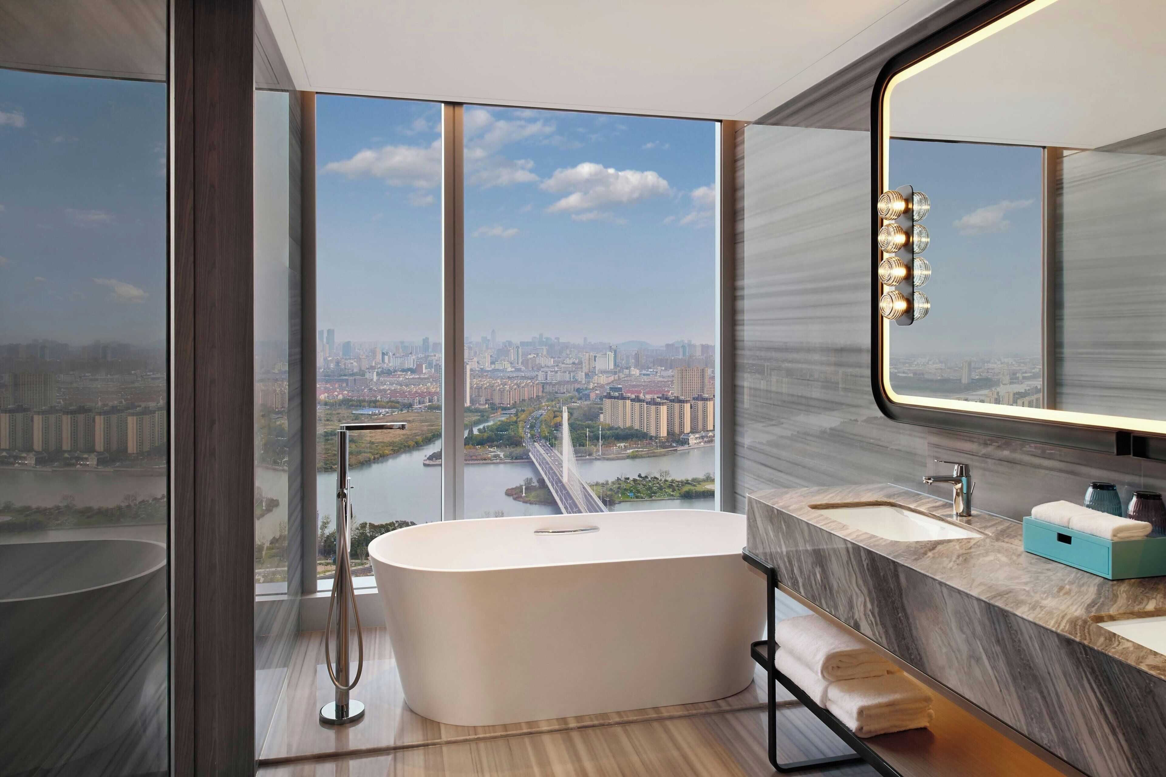 A modern bathroom with a large bathtub, a sink, and a mirror. It features a city view through large windows.