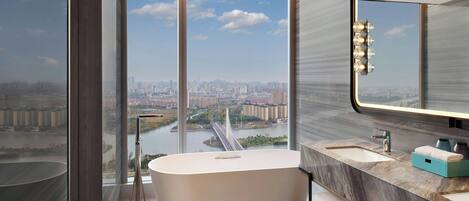 A modern bathroom with a large bathtub, a sink, and a mirror. It features a city view through large windows.