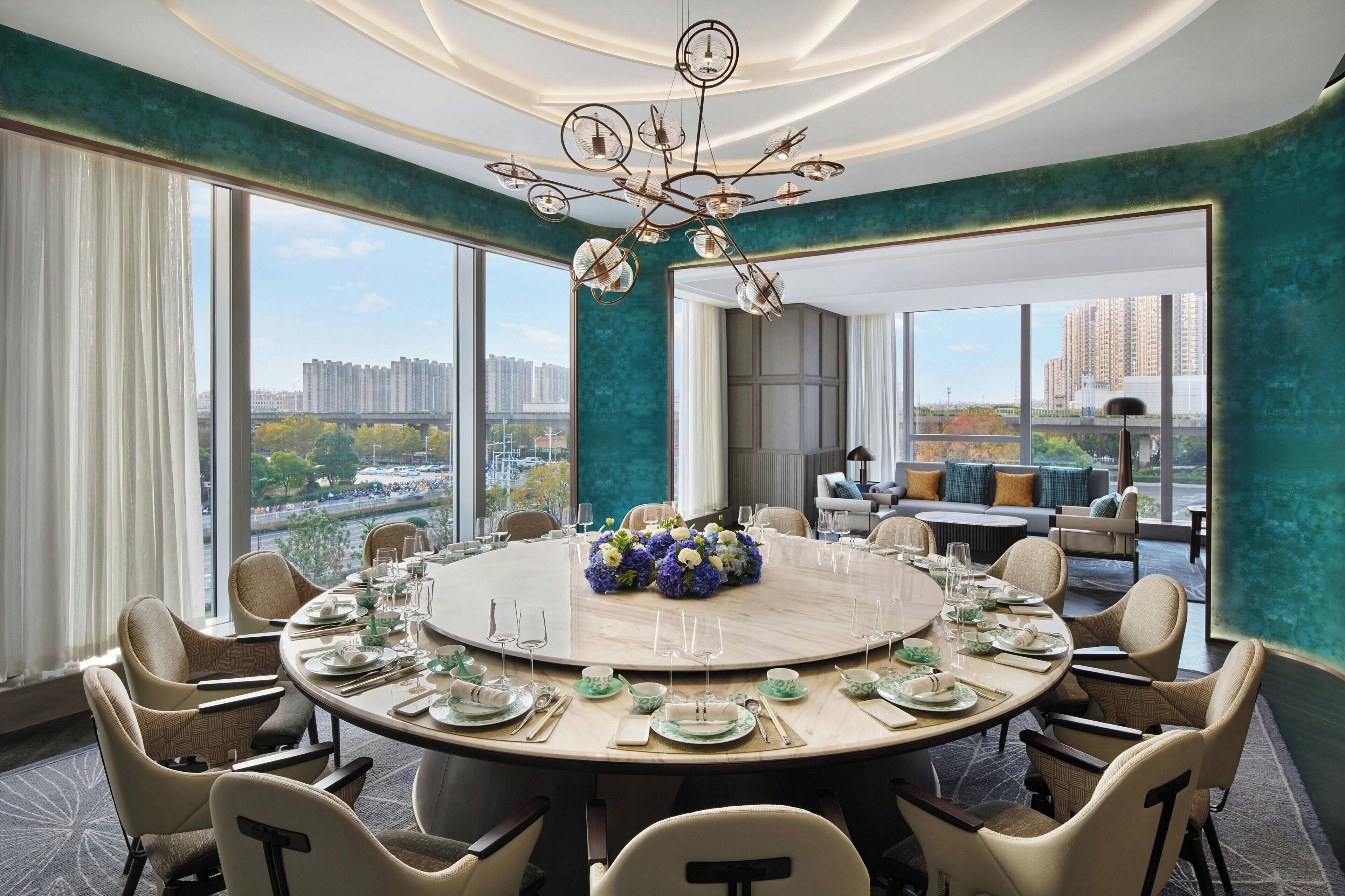 Une table de salle à manger ronde, mise pour un repas, avec des chaises, un lustre et une vue sur le paysage urbain à travers de grandes fenêtres.