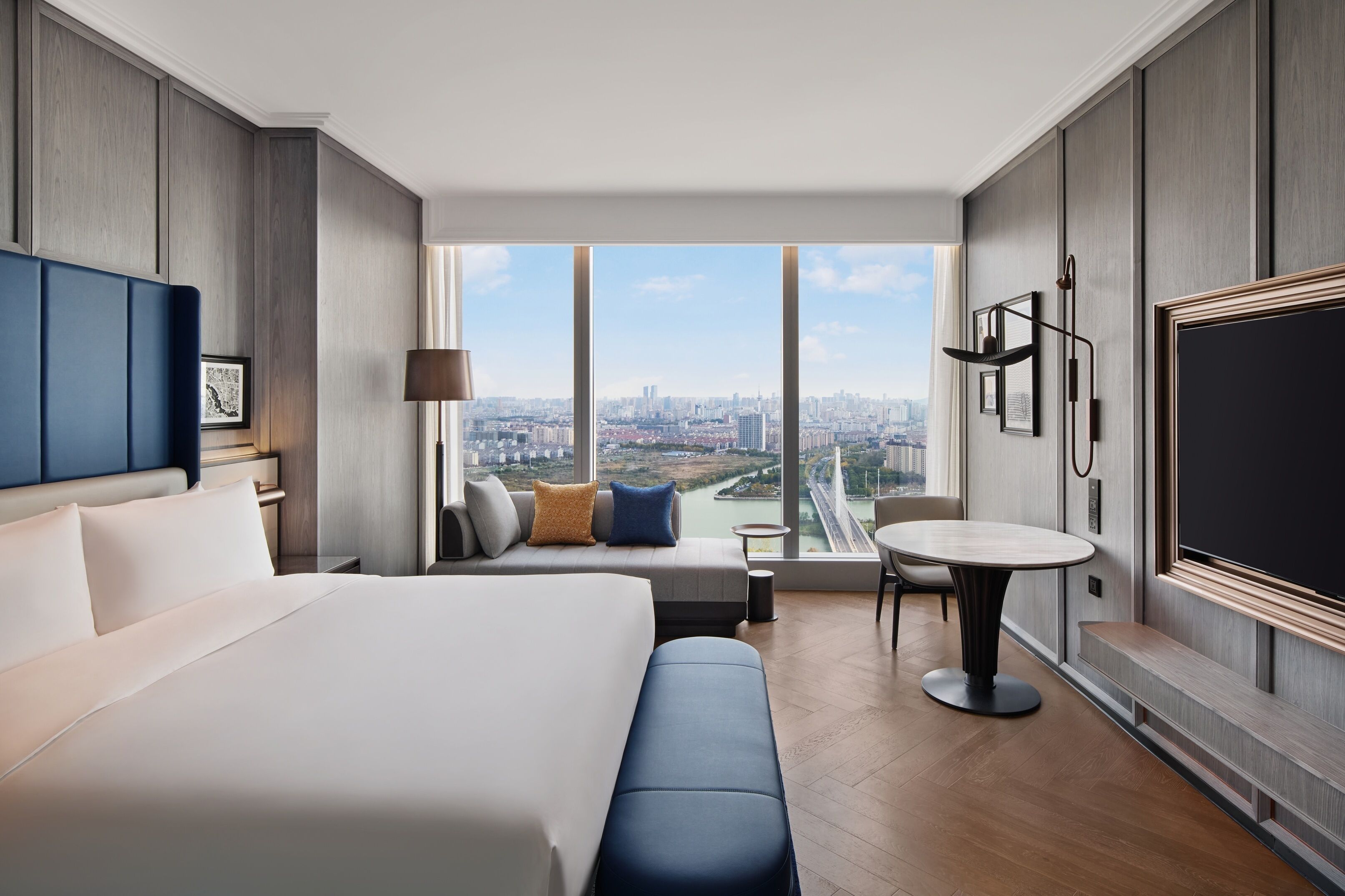 Ein modernes Hotelzimmer mit einem großen Bett, einer Sitzecke, einem Esstisch und Blick auf die Stadt durch große Fenster.